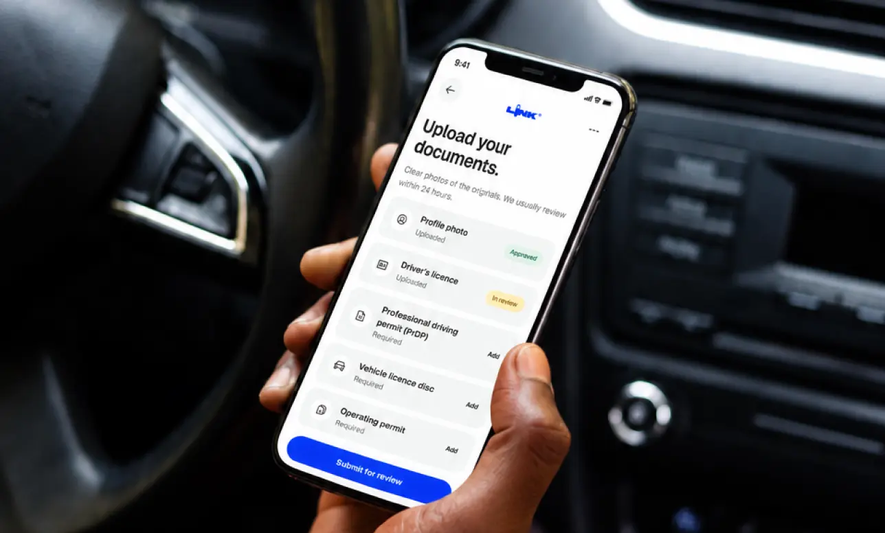 A hand holding a phone that shows the Link Driver app’s Upload your documents screen, inside a car